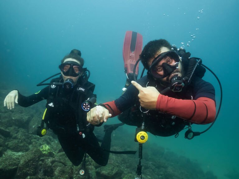 Two scuba divers underwater holding hands, one pointing at the other, with rocky terrain in the background, featured in the blog post ‘PADI Certifications, Why Get your Scuba diving PADI license in Costa Rica?’